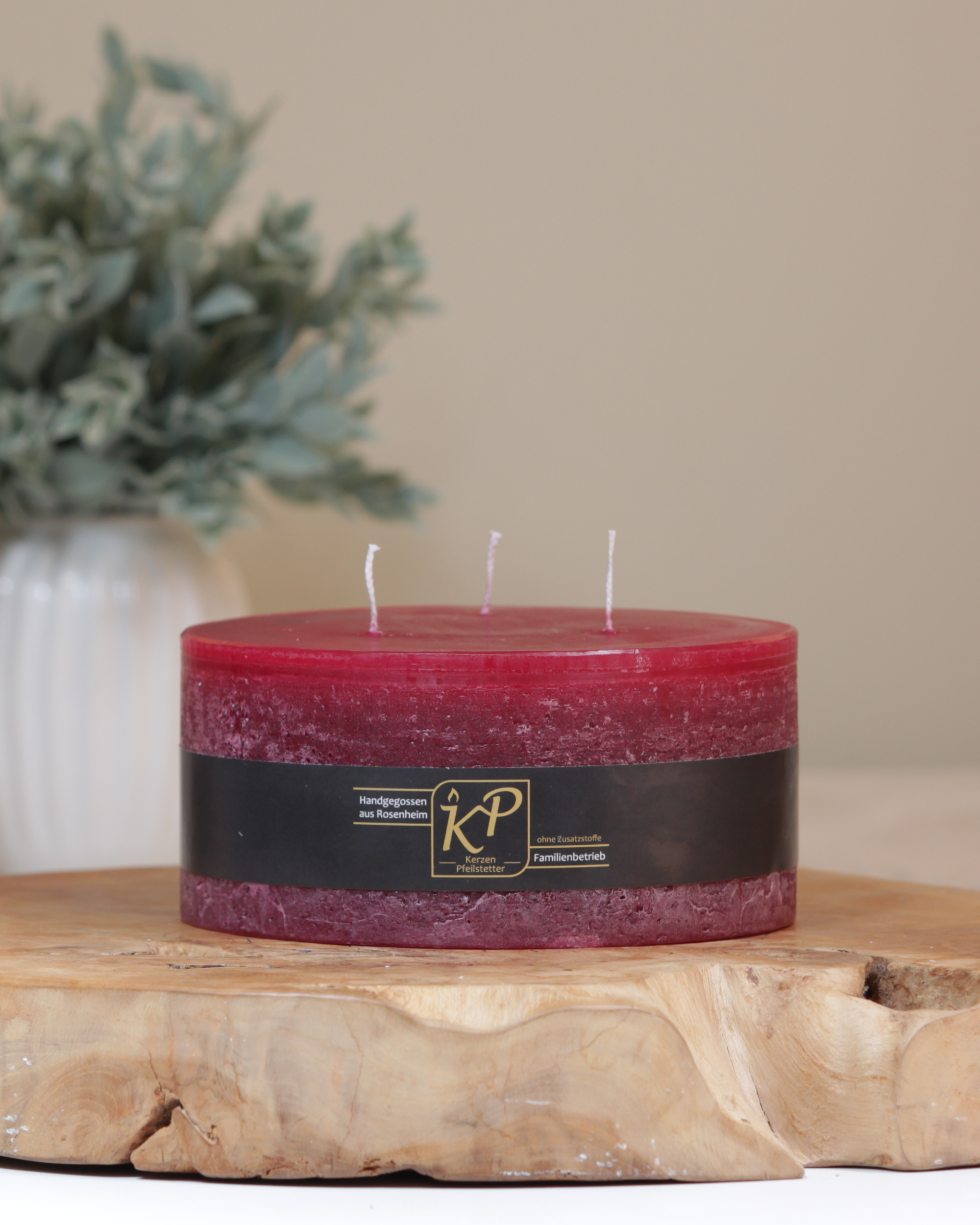 Dreidochtkerze in burgundrot dekorativ auf einem Holztisch mit Pflanze im Hintergrund.