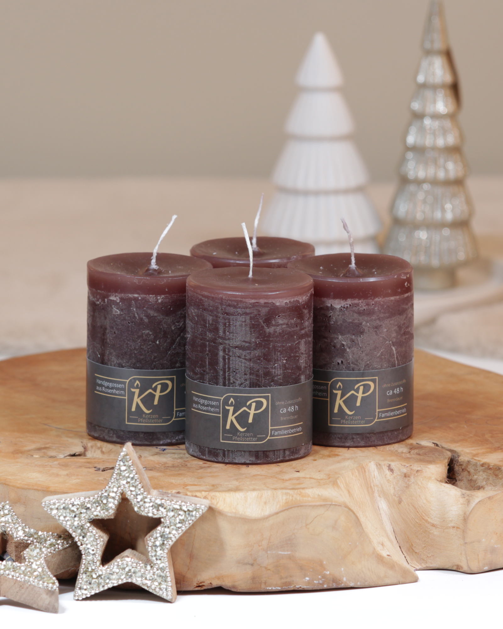 Vier Tischkerzen in dunkelbraun dekorativ auf einem Holztisch mit Weihnachtsdeko im Hintergrund.