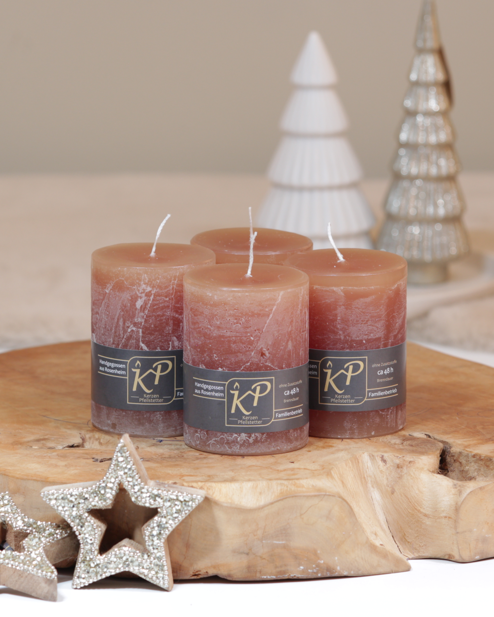 Vier Tischkerzen in karamellbraun dekorativ auf einem Holztisch mit Weihnachtsdeko im Hintergrund.