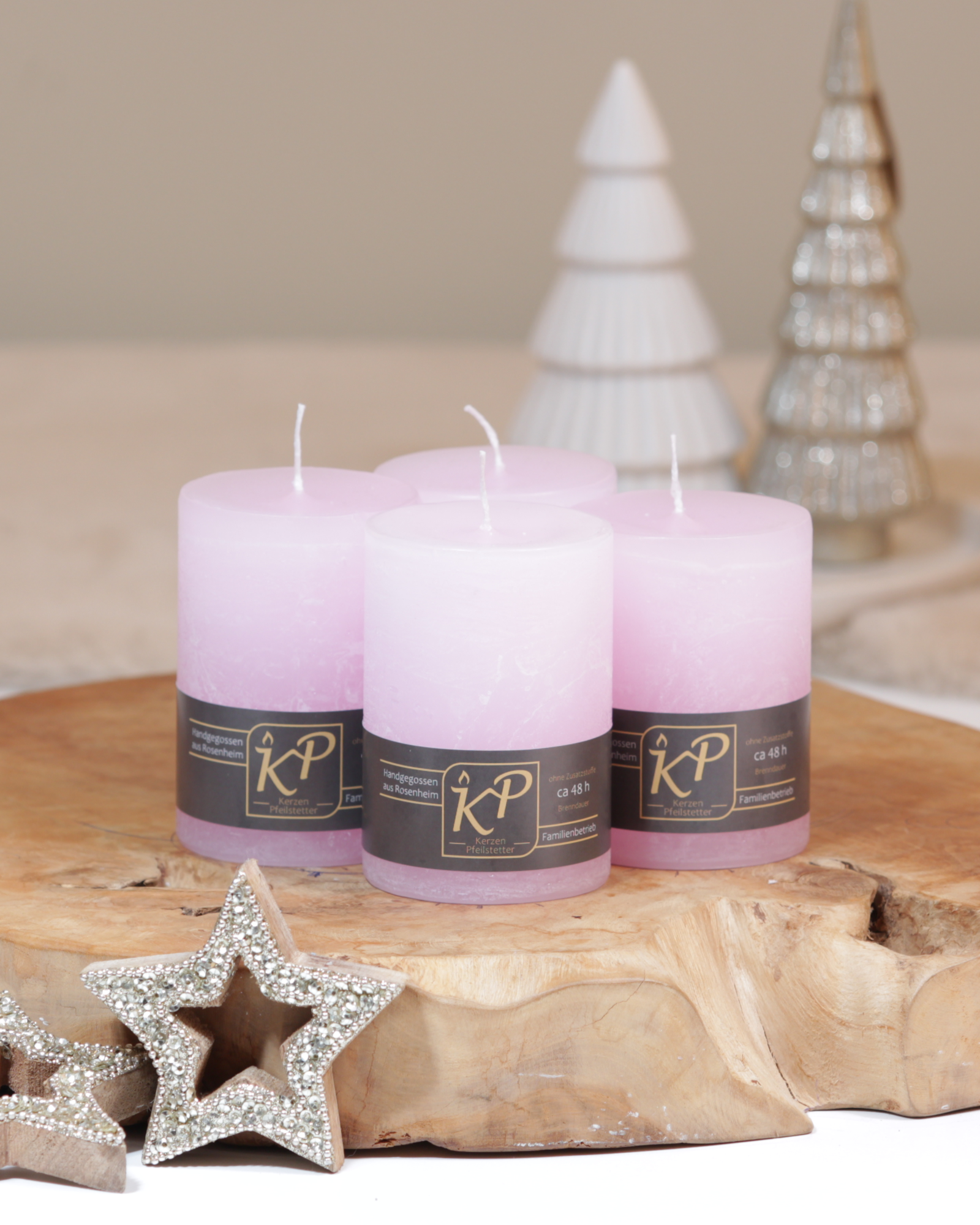 Vier Tischkerzen in pastellrosa dekorativ auf einem Holztisch mit Weihnachtsdeko im Hintergrund.