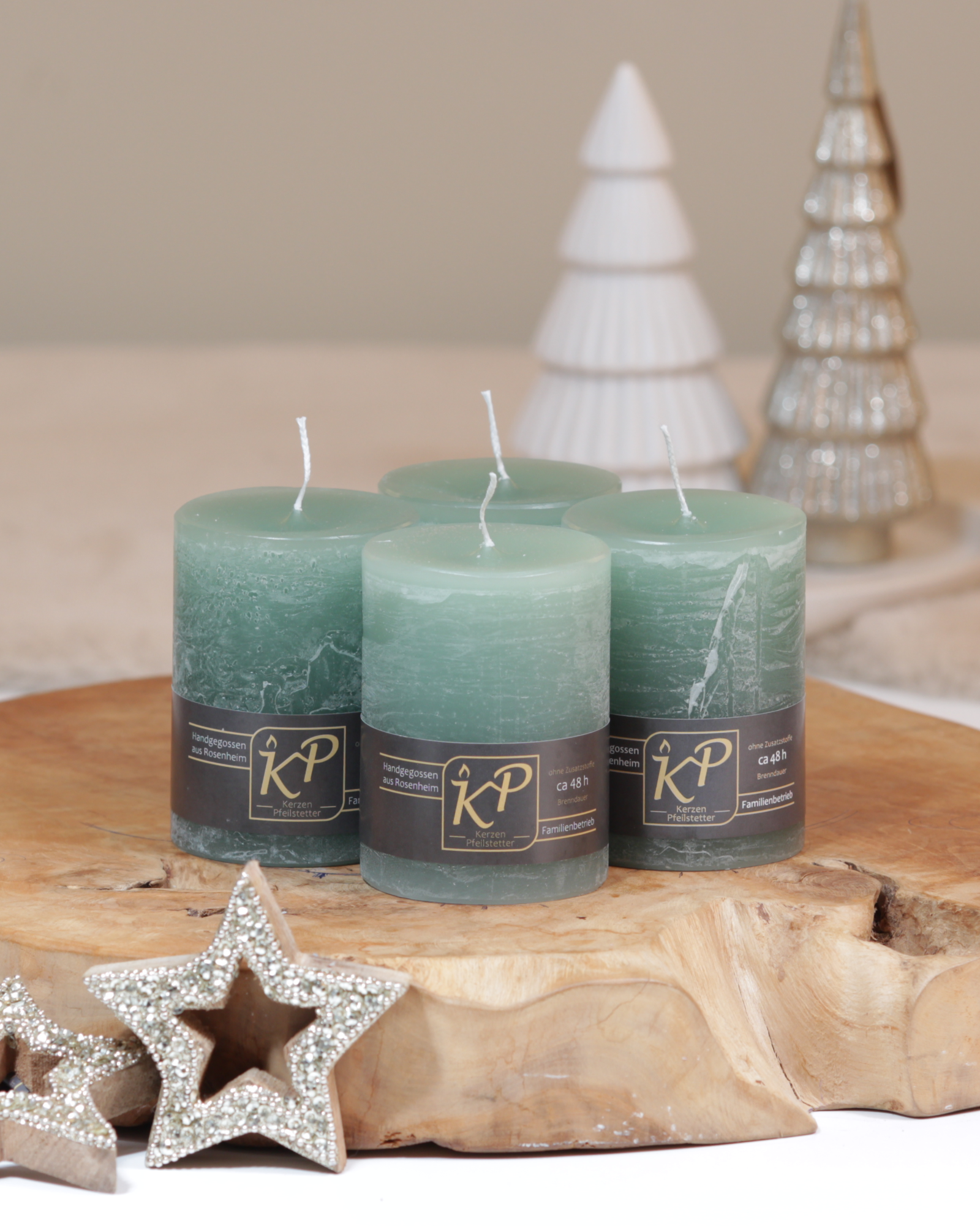 Vier Tischkerzen in salbeigrün dekorativ auf einem Holztisch mit Weihnachtsdeko im Hintergrund.