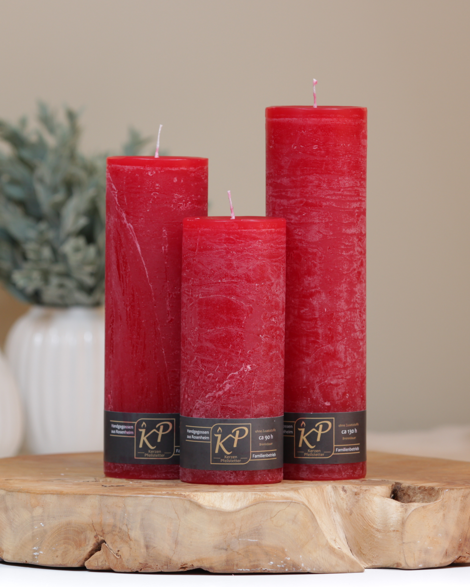 Blütenkerzen in weihnachtsrot in drei Größen – dekorativ auf einem Holztisch mit Pflanze im Hintergrund.