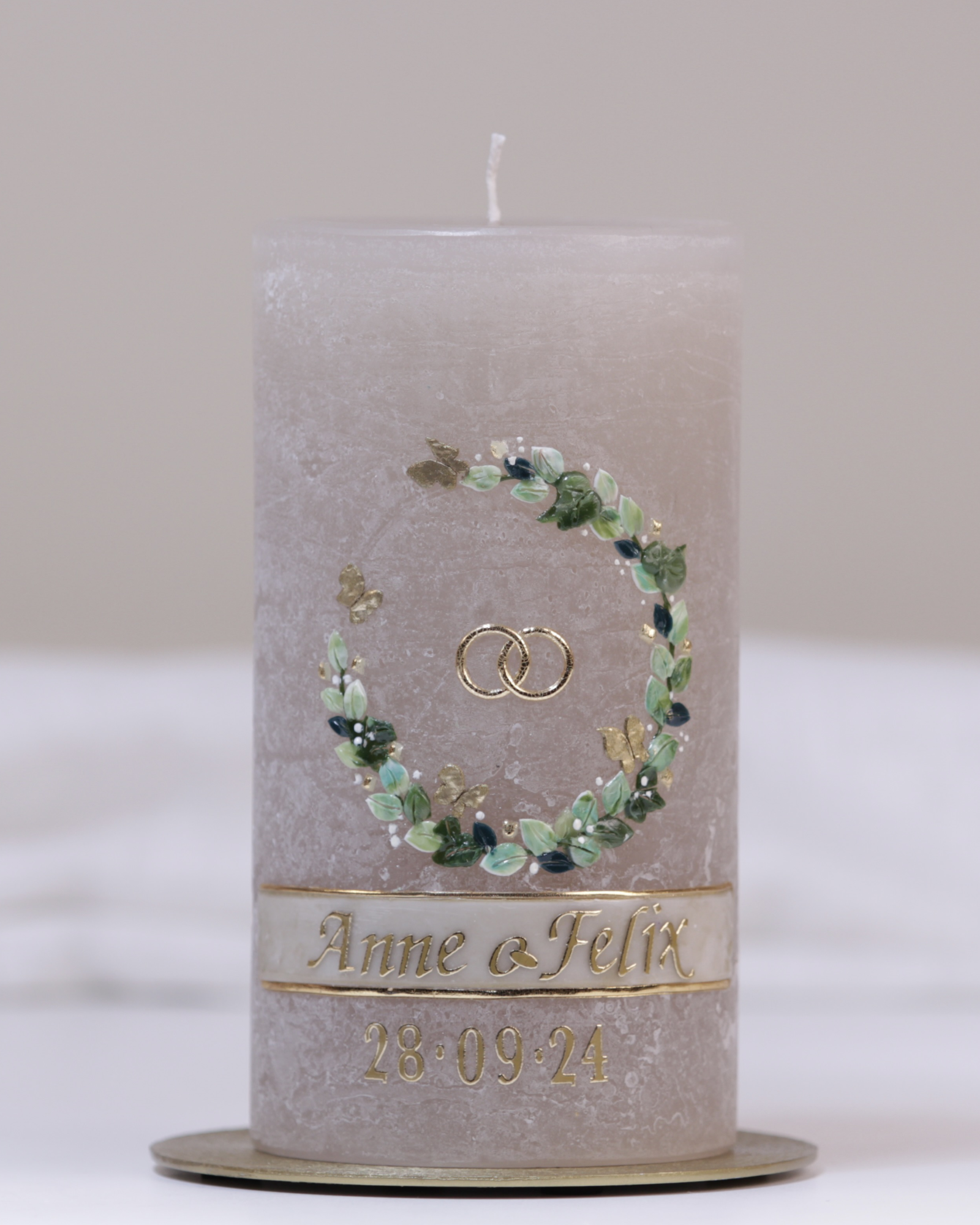 Sandgraue Hochzeitskerze mit Eheringen, grünem Blumenkranz und goldenen Schmetterlingen – detailverliebt gestaltet.