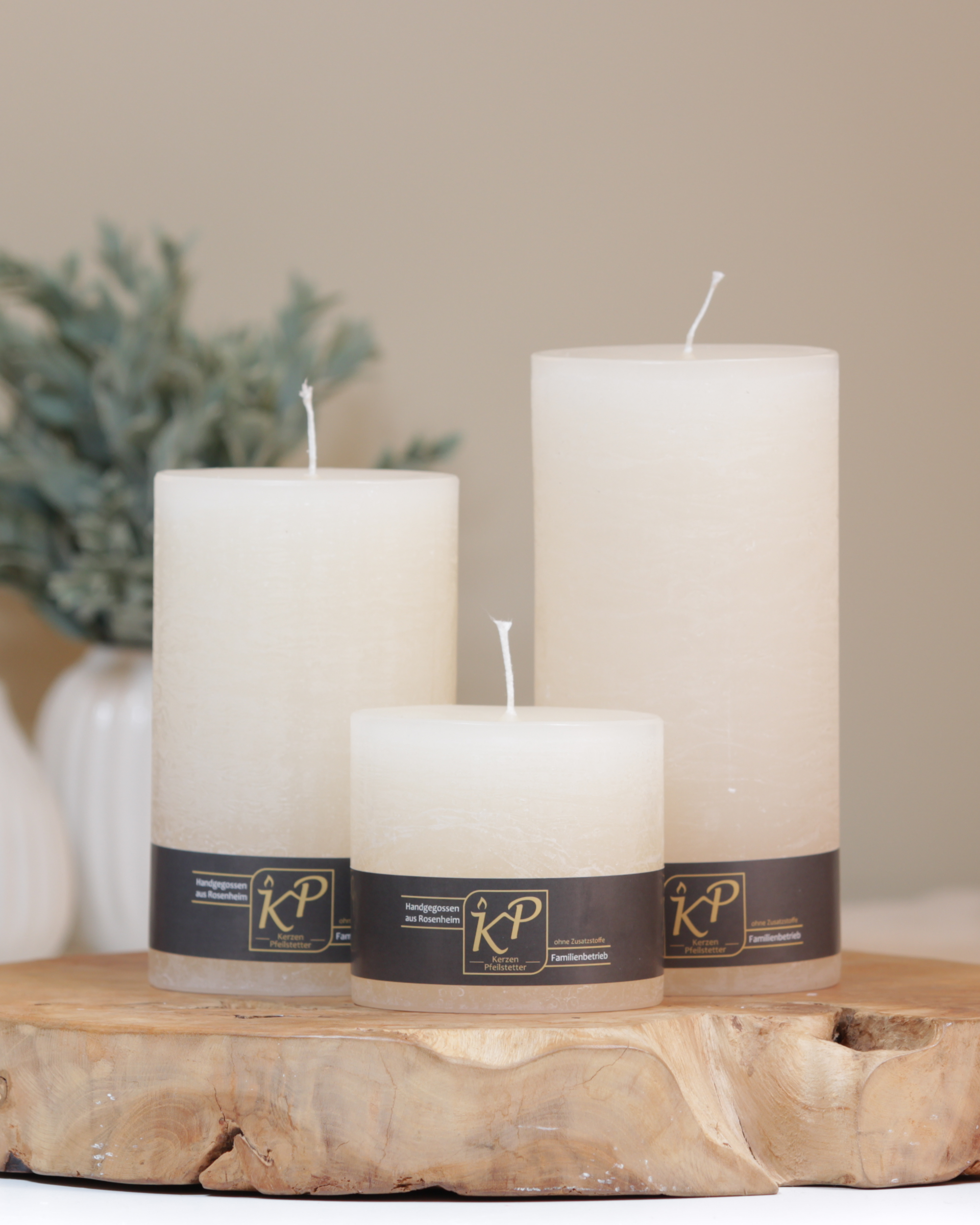Stumpenkerzen in creme in drei Größen – dekorativ auf einem Holztisch mit Pflanze im Hintergrund.