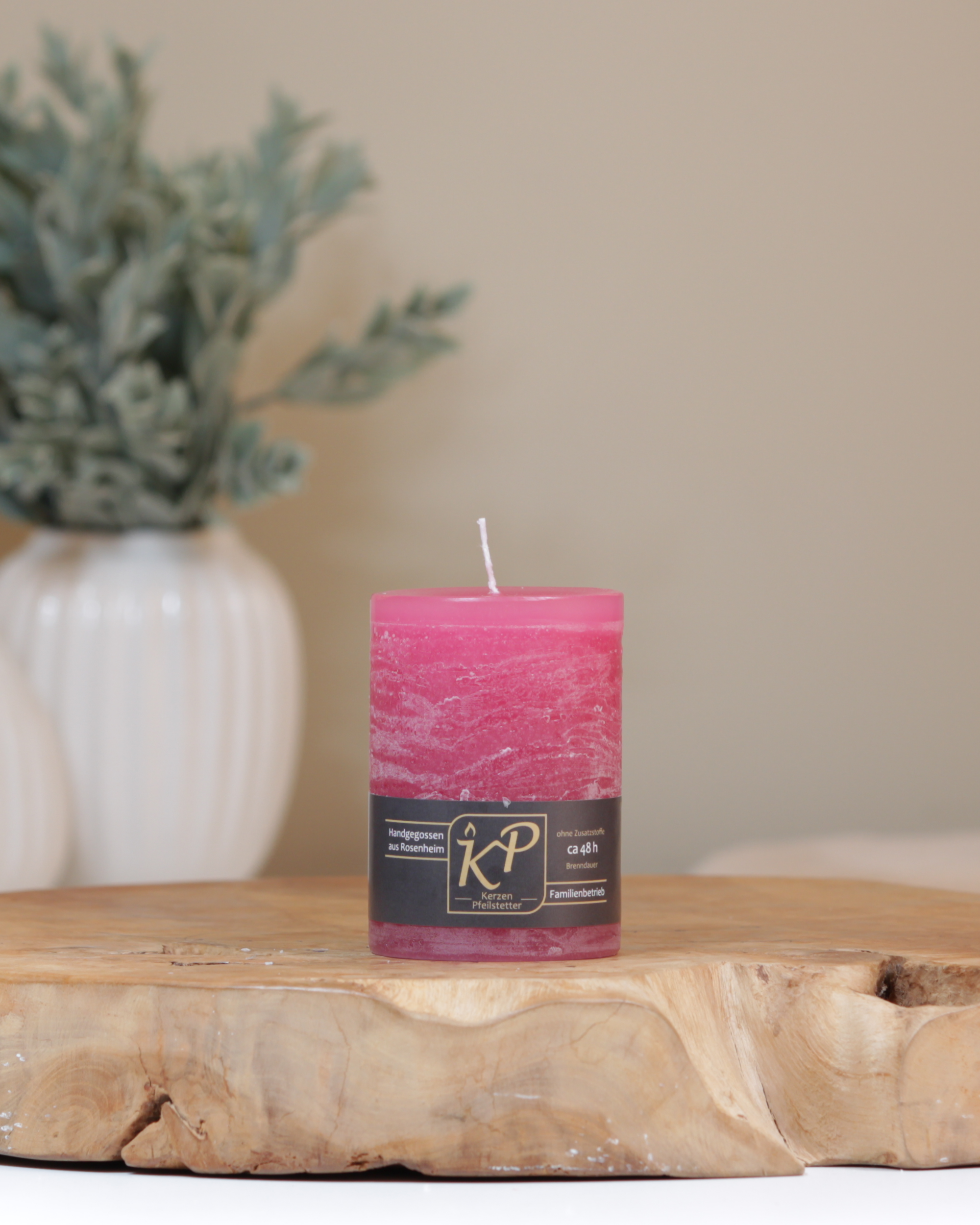 Tischkerze in himbeerrosa dekorativ auf einem Holztisch mit Pflanze im Hintergrund.