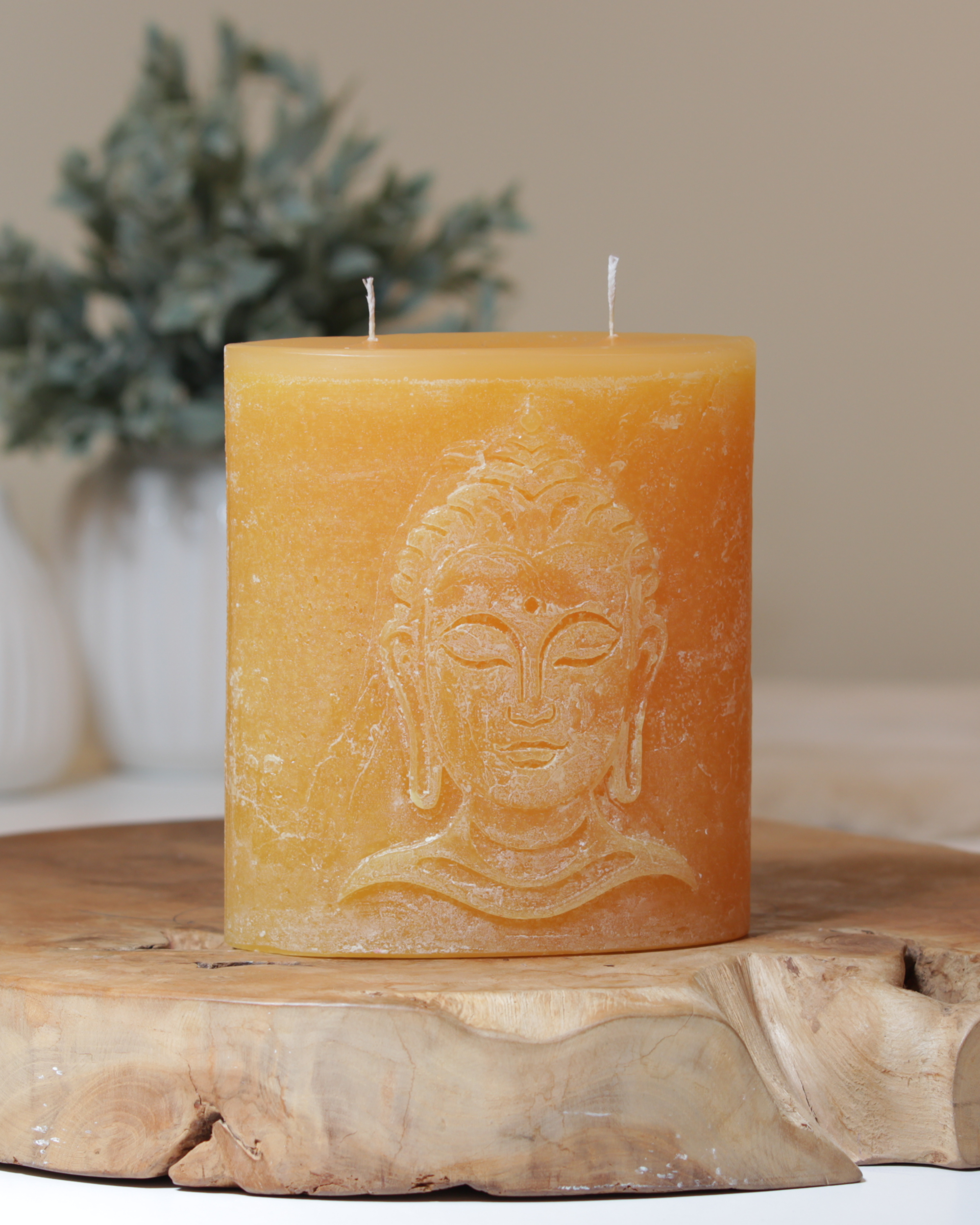 Zweidochtkerze mit Buddha-Motiv in sonnengelb dekorativ auf einem Holztisch mit Pflanze im Hintergrund.