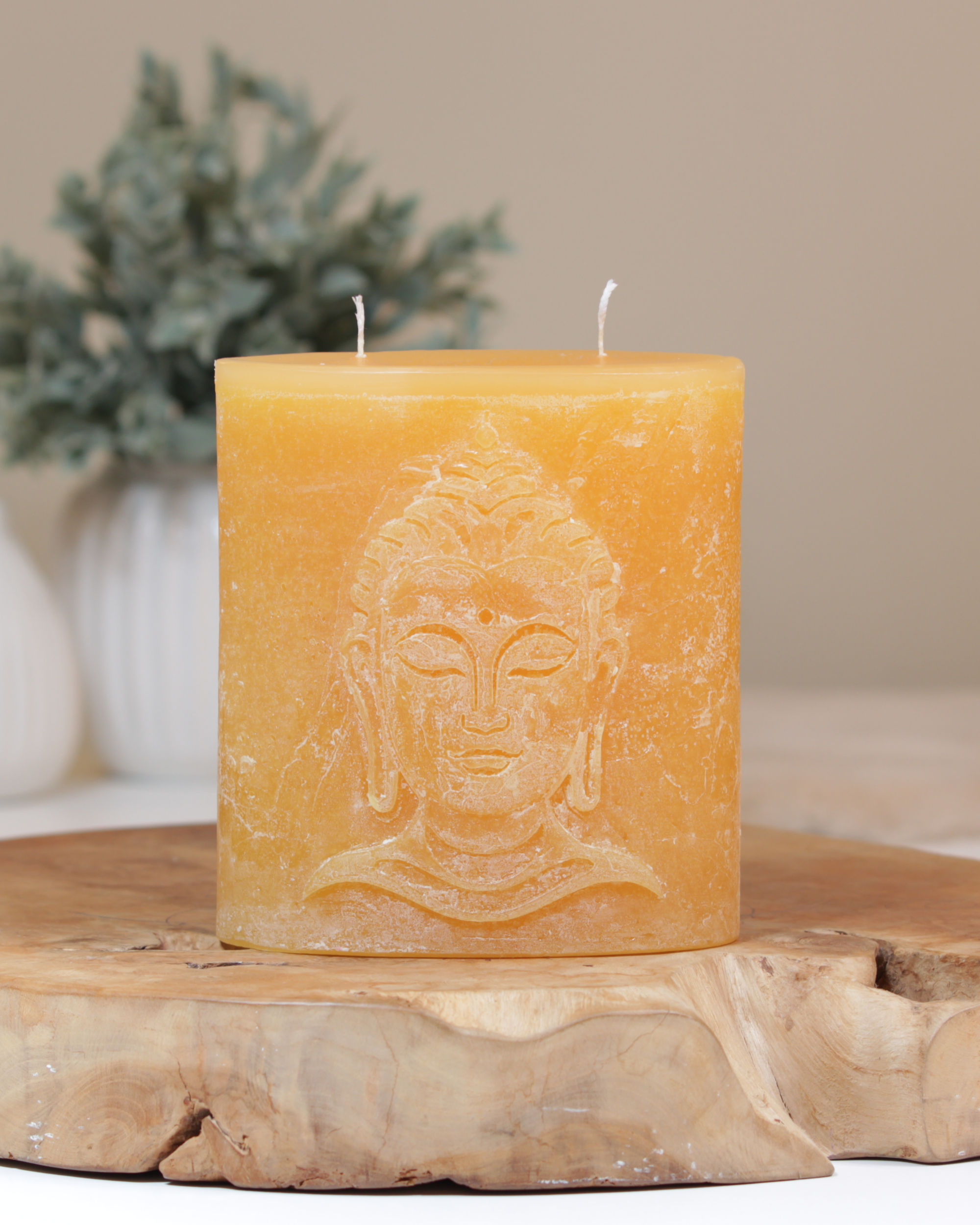 Zweidochtkerze mit Buddha-Motiv in sonnengelb dekorativ auf einem Holztisch mit Pflanze im Hintergrund.